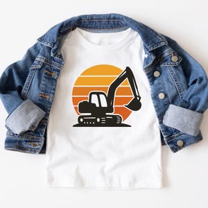 May include: White t-shirt featuring a black excavator graphic set against a gradient orange and yellow sunset design. The shirt is displayed on top of a denim jacket.