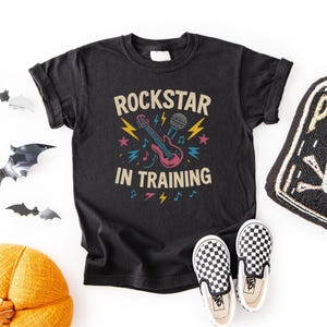 May include: Black t-shirt with the text "ROCKSTAR IN TRAINING" and a graphic of a guitar, microphone, and musical notes. The shirt is paired with black and white checkered slip-on shoes.