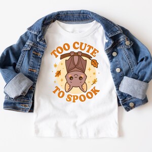 May include: White t-shirt with a bat graphic and the text "Too Cute to Spook" in orange. The bat is brown and hanging upside down from a branch. The shirt is paired with a blue denim jacket.