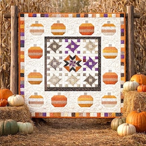 Op de afbeelding: Een herfstquilt met pompoen- en sterpatronen. De quilt heeft een witte achtergrond met oranje, bruine en paarse accenten. Pompoenen zijn rond de rand gerangschikt en sterren in het midden. De quilt wordt buiten tentoongesteld met pompoenen en hooibalen.