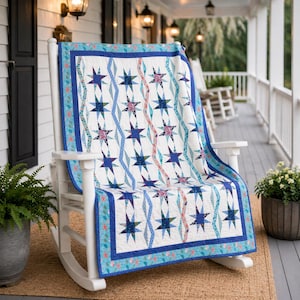 Puede incluir: Una mecedora blanca sostiene un edredón con estampado de estrellas. El edredón presenta estrellas azules, patrones de diamantes y un borde con detalles florales. La paleta de colores incluye tonos de azul, rosa y blanco. El edredón está sobre la silla en un porche.