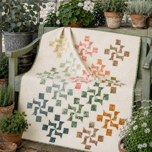 May include: A cream-coloured quilt featuring a repeating pinwheel pattern in a gradient of colours including yellow, orange, red, green, and blue. The fabric has a textured appearance, and the quilt is draped over a wooden bench.