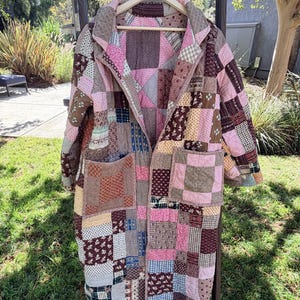 May include: A long-sleeved quilted coat featuring a patchwork design composed of various fabric squares in shades of pink, brown, blue, and cream. The coat includes a wide collar, two large front patch pockets, and is displayed on a wooden hanger.