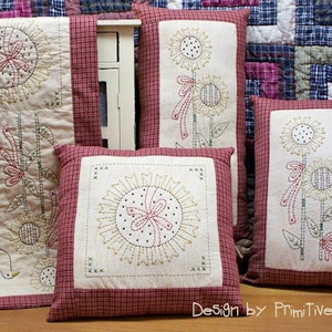 May include: A set of four decorative cushions with a red and white checked border. The cushions feature embroidered sunflowers with a bird and a bow. The cushions are made of a soft, white fabric and have a rustic, farmhouse style. "Design by Primitive Stitches" is printed on the bottom of the image.