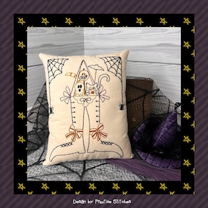 May include: A white cushion with an embroidered design of a house with a cat, ghost, and pumpkin inside. The house is made of legs and has bows on the legs. The cushion is on a black and white background with a purple hat.