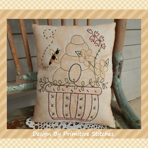 May include: A beige pillow with an embroidered design of a bee flying towards a beehive in a basket with flowers. The pillow is sitting on a wooden chair with a crocheted doily underneath. The text "Design By Primitive Stitches" is at the bottom of the image.