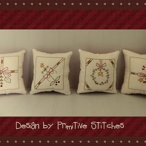May include: Four white throw pillows with embroidered Christmas designs. The pillows feature a bow, a wreath, a present, and a sprig of holly. The pillows are arranged in a row on a white surface. The text "Design by Primitive Stitches" is printed below the pillows.