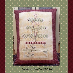 Puede incluir: Una almohada beige con un borde rojo presenta un diseño bordado de un árbol de Navidad estilizado con corazones rojos y un lazo. La almohada está sobre una superficie blanca con un patrón de cuadros rojos y blancos. El texto "Design by Primitive Stitches" está en la parte inferior de la imagen.