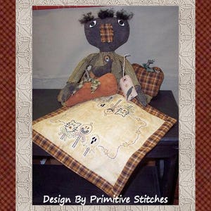 May include: A black rag doll with a plaid dress and a pumpkin in its arms sits on a pillow with a Halloween-themed embroidery design. The pillow has a brown and tan plaid border. The text "Design By Primitive Stitches" is at the bottom of the image.