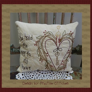 May include: Cream-colored decorative pillow with embroidered heart and key design. The heart is outlined in brown thread and adorned with leaves and red berries. Text reads "You hold the key to my heart." The pillow is on a wooden chair.