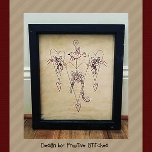Peut inclure: &OElig;uvre d'art encadr&eacute;e avec un motif cousu de trois c&oelig;urs, de fleurs et d'un oiseau. L'&oelig;uvre est dans un cadre noir et a un fond beige. Le texte "Design by Primitive STITCHES" est en bas.