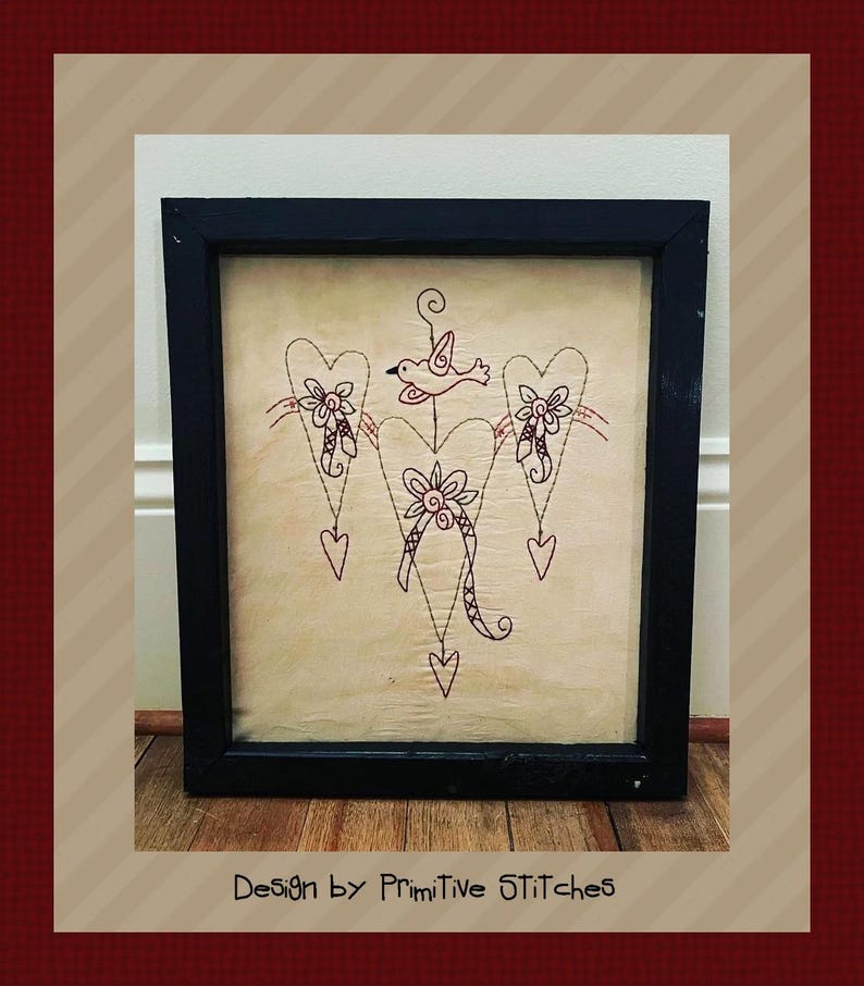 Peut inclure: &OElig;uvre d'art encadr&eacute;e avec un motif cousu de trois c&oelig;urs, de fleurs et d'un oiseau. L'&oelig;uvre est dans un cadre noir et a un fond beige. Le texte "Design by Primitive STITCHES" est en bas.