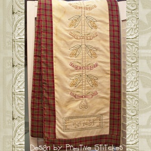 May include: A beige table runner with a red and green plaid border. The runner is decorated with embroidered fall-themed designs, including corn stalks, bows, and the word "Harvest".