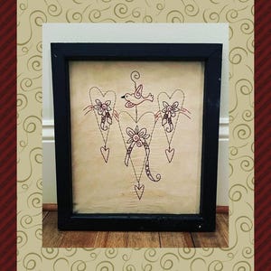 Peut inclure: Une &oelig;uvre d'art encadr&eacute;e avec un motif cousu de c&oelig;urs, de fleurs et d'un oiseau dans des tons bruns et cr&egrave;me. L'&oelig;uvre est dans un cadre noir et est expos&eacute;e sur une surface en bois. Le texte "Design by Primitive Stitches" est en bas.