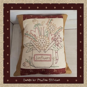 May include: A cream-colored pillow with a brown border features an embroidered design of sunflowers, hearts, and other flowers in a bucket. The bucket has a label that reads "Sunflowers". The pillow is decorated with a brown and red fabric border.
