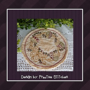 Puede incluir: Un posavasos redondo de tela beige con un diseño bordado de una corona de hojas verdes, bayas rojas y huevos amarillos y marrones. El texto "Design by Primitive Stitches" está debajo del posavasos.