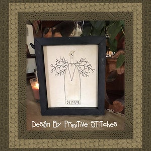 Könnte beinhalten: Ein gerahmtes Stickbild mit einem stilisierten Engel mit Flügeln aus Ästen und dem darunter gestickten Wort "BELIEVE". Das Design ist in einem schwarzen Rahmen und befindet sich auf einer braunen Oberfläche.