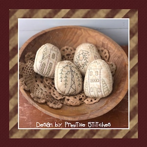 May include: Four handmade fabric ornaments in the shape of houses and a tree, displayed in a wooden bowl. The ornaments are beige with brown stitching and feature simple designs. The text "Design by Primitive Stitches" is visible at the bottom of the image.