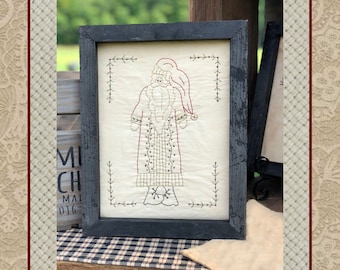 May include: A framed cross-stitch design of a red and white Santa Claus with a black beard and a green hat. The design is on a white background and is surrounded by a green border. The frame is black with a distressed finish. The text "Design by Primitive Stitches" is below the frame.