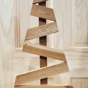 Può includere: Una scultura a forma di albero di Natale in legno. L'albero è costruito con assi di legno marrone chiaro, disposte a zig-zag attorno a un supporto verticale centrale. Anche la base è in legno.