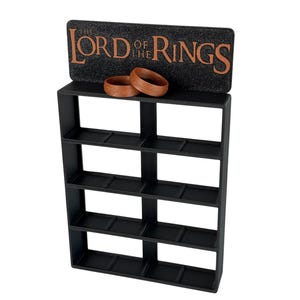 Peut inclure: Un présentoir rectangulaire noir doté de quatre étagères horizontales et d'un panneau supérieur portant l'inscription "THE LORD OF THE RINGS" en lettres cuivrées. Deux anneaux marron avec une écriture gravée reposent sur l'étagère supérieure.