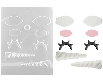 Op de afbeelding: Een transparante plastic mal met verschillende vormen met een eenhoorn-thema, waaronder ogen, hoorns en wimpers. De mal wordt getoond met afgewerkte chocolade stukken in wit, roze en zwart.