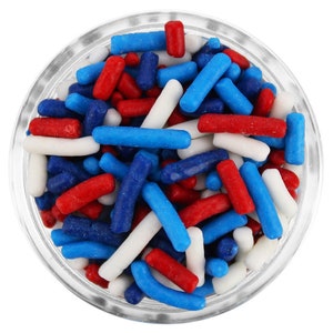 May include: A clear glass bowl filled with red, white, and blue sprinkles. The sprinkles are cylindrical and of varying lengths, creating a colorful and festive appearance. The bowl is set against a white background.