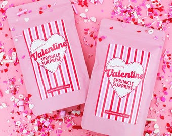 May include: Two pink bags of Valentine sprinkles with a heart-shaped label that reads "Valentine Sprinkle Surprise." The bags are surrounded by scattered sprinkles in various shapes and colors, including hearts and rods. Each bag contains 4 ounces (113 grams) of sprinkles.