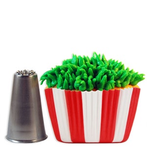 May include: A silver metal piping tip with a star-shaped opening, next to a red and white striped cupcake liner with green frosting piped to look like grass.