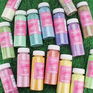 Op de afbeelding: Een verzameling doorzichtige plastic flessen gevuld met kleurrijke sprinkles. Elke fles heeft een witte deksel en een roze label met de tekst "Sprinkles!" en een cupcake-logo. De sprinkles zijn er in verschillende kleuren, waaronder groen, blauw, roze, geel en bruin.