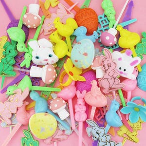 May include: A collection of colorful Easter-themed cake toppers. The toppers feature various designs, including bunnies, chicks, eggs, flowers, and mushrooms. The toppers are in shades of pink, yellow, green, blue, and white, with gold accents. The background is a solid pink.