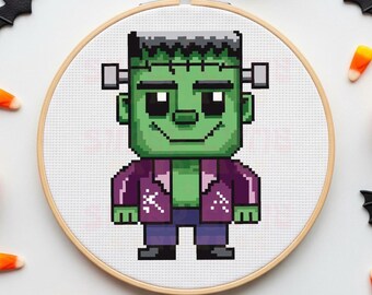 Puede incluir: Un bastidor de bordado de punto de cruz con un monstruo de Frankenstein pixelado. El monstruo tiene piel verde, cabello negro y lleva una chaqueta morada y pantalones azules. El fondo es blanco y el bastidor es de madera. Tema de Halloween.