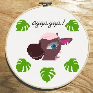 May include: A white embroidery hoop with a pink and brown cartoon creature with the text "awww yeah!" and green monstera leaves around the creature.