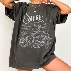 May include: A charcoal gray oversized t-shirt featuring a white line art graphic of a hobbit hole, a smoking pipe, a book, and a teacup. The text reads THE SHIRE and All we have to decide is what to do with the time that is given to us.