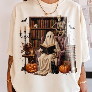 Op de afbeelding: Een gebroken wit t-shirt met een grafische print van een geest in een laken die een boek leest in een fauteuil, een skelet, een zwarte kat, pompoenen, een kandelaar en een boekenplank. Het ontwerp bevat vleermuizen en schedels in een scène.