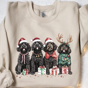 May include: A beige sweatshirt with a watercolour illustration of four black dogs. Each dog is dressed for Christmas, wearing Santa hats, reindeer antlers, and festive sweaters. They are sitting in front of wrapped gifts.