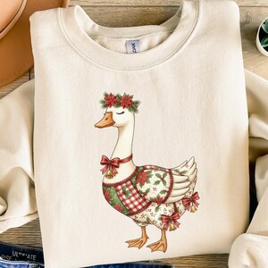 May include: Cream-colored sweatshirt featuring a whimsical goose design. The goose is adorned with a floral crown, red bows, and a festive patterned vest. The sweatshirt is paired with blue denim shorts, a tan hat, and sandals.
