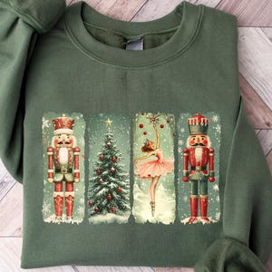 May include: An olive green sweatshirt featuring a festive design. The design includes a Christmas tree, a ballerina, and two nutcrackers in red and green. The sweatshirt is made of a soft material and is perfect for the holiday season.