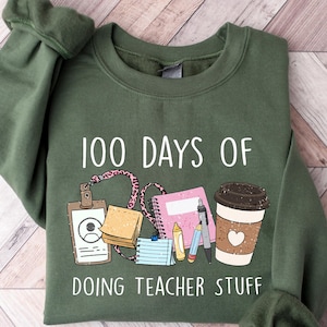 May include: A green sweatshirt featuring the phrase "100 Days of Doing Teacher Stuff" alongside illustrations of school essentials, such as a coffee cup, notepad, and pencils. The garment is made of a soft fabric.