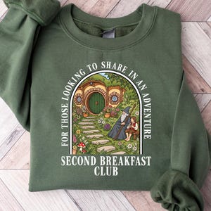 May include: An olive green crewneck sweatshirt featuring a graphic print of a round hobbit-style door, a wizard, and a small figure sitting on a bench. The text reads: FOR THOSE LOOKING TO SHARE IN AN ADVENTURE SECOND BREAKFAST CLUB in white.