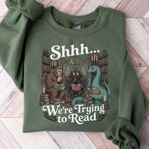 Può includere: Una felpa girocollo verde oliva con un'illustrazione grafica di Bigfoot, Mothman e il mostro di Loch Ness che leggono libri in una biblioteca. Il testo sopra e sotto l'illustrazione recita Shhh... We're Trying to Read in carattere bianco.