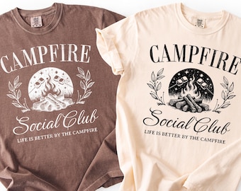 May include: Two t-shirts, one brown and one cream, with a camping-themed graphic. The design includes the words "Campfire Social Club" and "Life is better by the campfire" around a campfire illustration. The shirts are casual and comfortable.