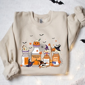 May include: Beige sweatshirt with a Halloween-themed graphic. The design features cartoon potion bottles with pumpkin, witch, and ghost characters. The bottles have labels like "Pumpkin Spice Proposal" and "Spiced Vanilla Versed". Black bats and orange leaves are scattered around the design.