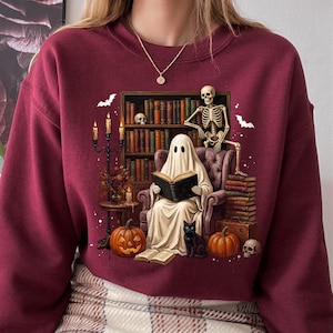 Könnte beinhalten: Ein bordeauxrotes Langarm-Sweatshirt mit einem Grafikdruck eines Geistes, der in einem Sessel sitzt und ein Buch liest, umgeben von einem Skelett, einer schwarzen Katze, Kürbissen, einem Kandelaber und einem mit Büchern gefüllten Regal.