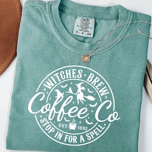 Puede incluir: Una camiseta de manga corta color verde salvia con un gráfico circular blanco que dice Witches Brew Coffee Co. Stop In For A Spell Est. 1692. El diseño incluye una bruja en una escoba, un gato, murciélagos, estrellas y un caldero.