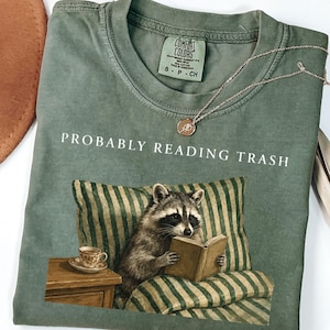 Op de afbeelding: Een saliegroen t-shirt met ronde hals en een afbeelding van een wasbeer die een boek leest op een gestreept kussen. De tekst "PROBABLY READING TRASH" staat in het wit boven de wasbeer. Een klein theekopje staat op een houten bijzettafeltje.