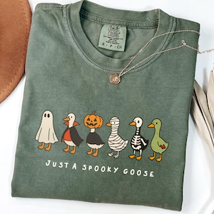 Puede incluir: Una camiseta verde oliva con un gráfico de seis gansos de dibujos animados disfrazados para Halloween. El texto "JUST A SPOOKY GOOSE" está impreso debajo. Un collar plateado con un pequeño colgante está en la camisa.