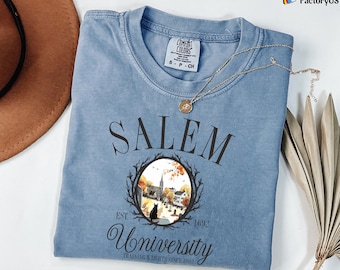 May include: A blue Comfort Colors t-shirt with the word "SALEM" printed across the chest. Below the word "SALEM" is a circular graphic with a black cat, church, and fall foliage. The text "EST. 1692" and "University" are also printed on the shirt.
