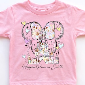 K&ouml;nnte beinhalten: Rosafarbenes T-Shirt mit kurzen &Auml;rmeln und einer Grafik eines Schlosses, Mickey Mouse-Ohren und Luftballons. Das Design enth&auml;lt den Text "Happiest place on Earth" in Schreibschrift. Die Grafik ist in Pastellfarben gehalten.