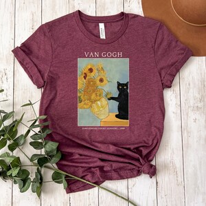 May include: Burgundy t-shirt featuring a print of Van Gogh's "Sunflowers" painting with a black cat. The text "VAN GOGH" is above the image, and "SUNFLOWERS (THIRD VERSION) 1888" is below. The shirt has short sleeves and a crew neck.